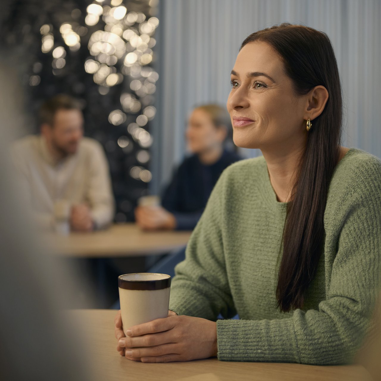 Eine Frau mit langem dunklem Haar sitzt an einem Tisch und hält einen Kaffeebecher. Im Hintergrund sitzen weitere Personen und unterhalten sich.
