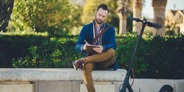 Ein Mann mit E-Roller sitzt draußen auf einer Bank und notiert etwas auf seinem Tablet