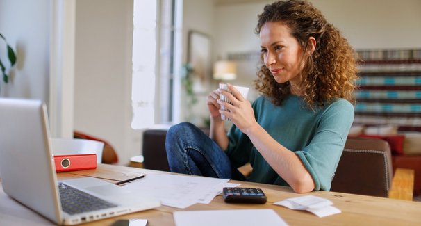 Frau sitzt entspannt mit Kaffee in der Hand am Schreibtsch mit Laptop