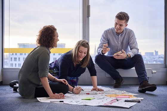 Drei Kolleginnen und Kollegen von Dataport sitzen zusammen, teilweise auf dem Boden und arbeiten an einem Mindmap.