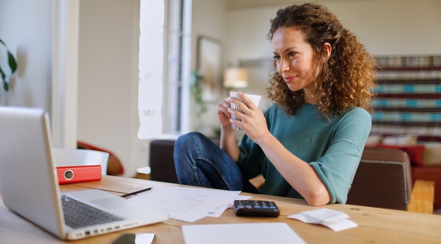 Frau sitzt entspannt mit Kaffee in der Hand am Schreibtsch mit Laptop