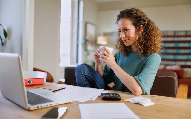 Frau sitzt entspannt mit Kaffee in der Hand am Schreibtsch mit Laptop