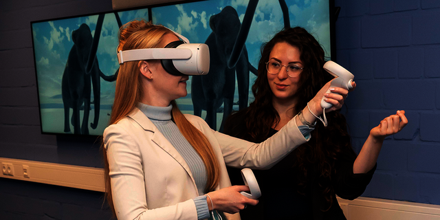 Eine Frau erklärt einer zweiten Frau mit VR-Brille die Funktionsweise von Virtual Reality