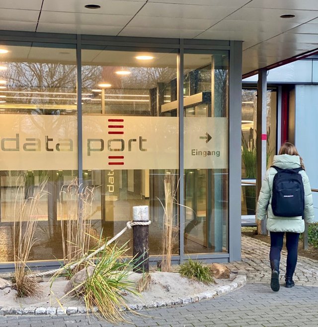 Eine Mitarbeiterin läuft mit einem Dataport Onboarding-Rucksack auf dem Rücken auf den Haupteingang des Dataport Standorts in Altenholz zu.