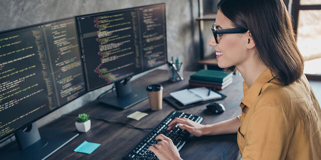 Eine Frau sitzt an einem Schreibtisch mit zwei Bildschirmen und tippt auf einer Tastatur.