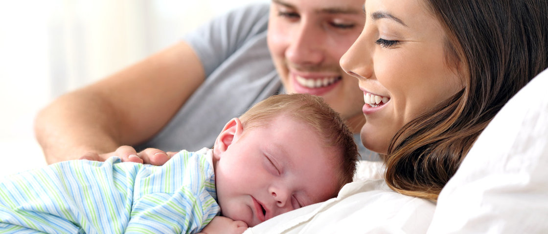 Ein junges Elternpaar hält im Bett ihr Baby auf dem Arm
