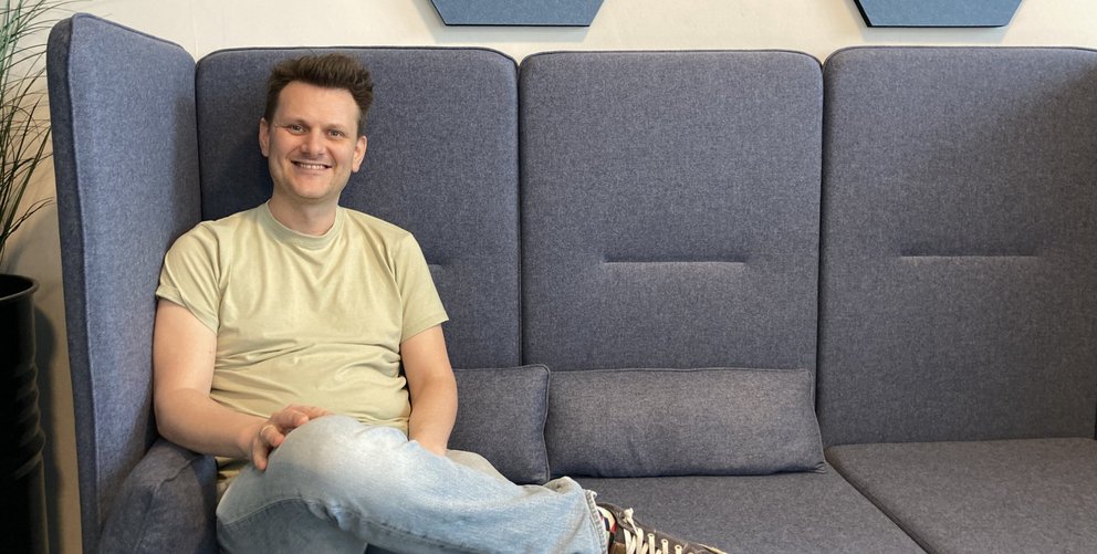 IT-Architekt Fabian Lange sitzt auf dem Sofa und lächelt freundlich in die Kamera.