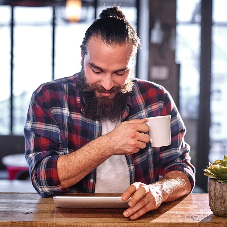 Ein Mann mit Dutt trinkt Kaffee und sieht aufs Tablet