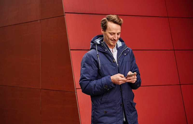 Mann mit Jacke vor einem Gebüde mit Handy in der Hand