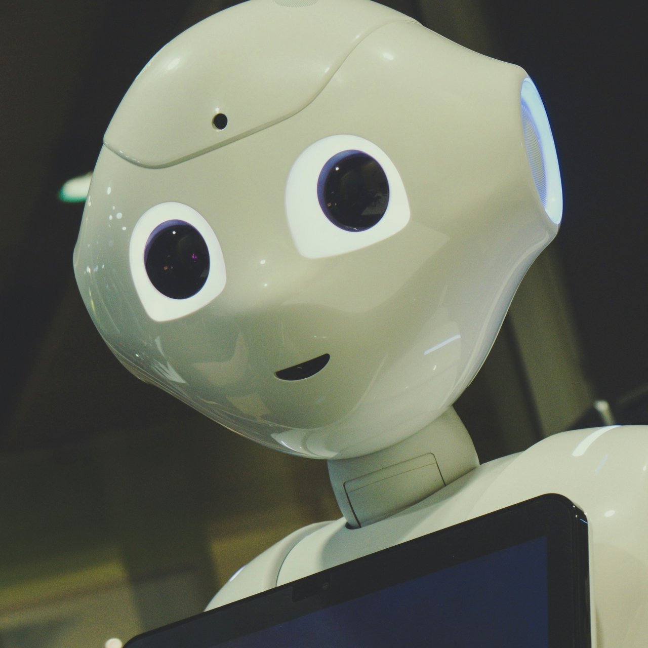 Ein humanoider Roboter mit großen Augen steht in einem Innenraum. Er hat ein Display auf der Brust.