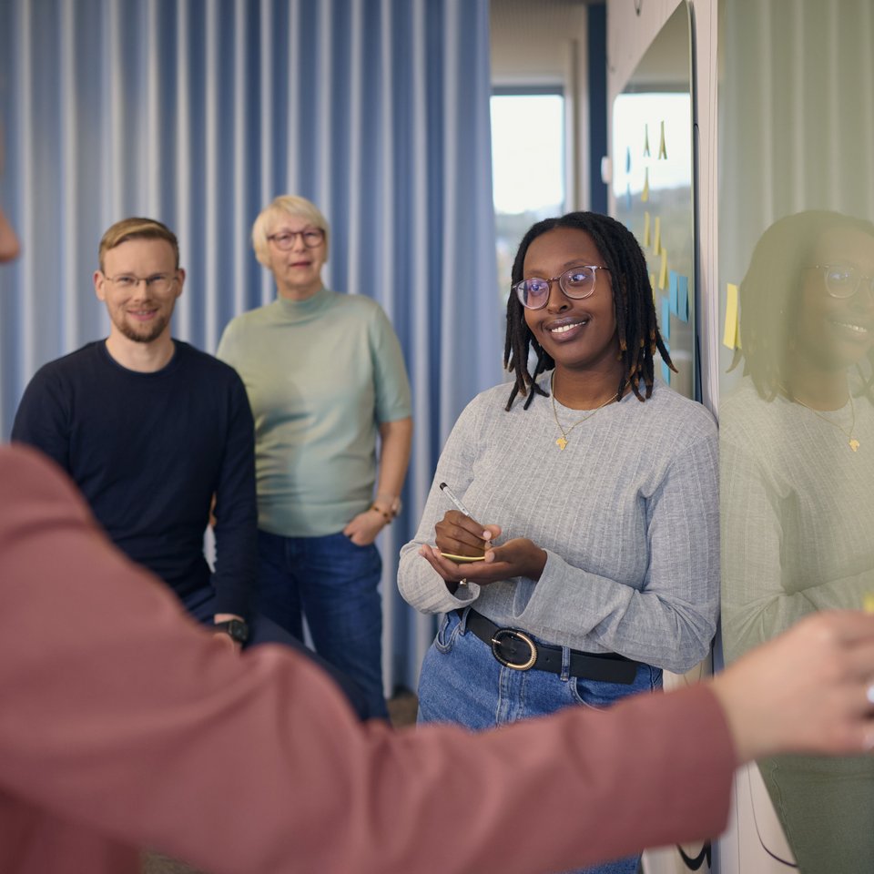 Vier Personen stehen vor einem Whiteboard mit Post-its. Eine Frau schreibt etwas auf, die anderen beobachten oder lachen.