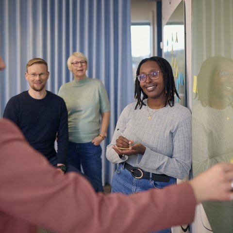 Vier Personen stehen vor einem Whiteboard mit Post-its. Eine Frau schreibt etwas auf, die anderen beobachten oder lachen.
