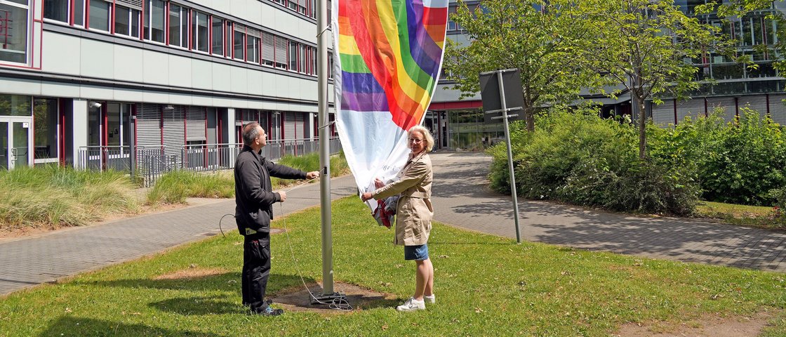 Zwei Menschen stehen neben einem Mast und hissen eine Regenbogenflagge