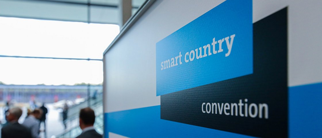 Menschen auf einer Messe. Im Vordergrund ist das Logo der Smart Country Convention zu sehen. 
