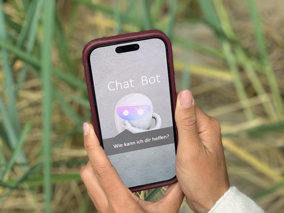 Ein Smartphone zeigt eine Chatbot-Anwendung auf dem Bildschirm. Das Gerät wird von zwei Händen gehalten, im Hintergrund sind Pflanzen zu sehen.