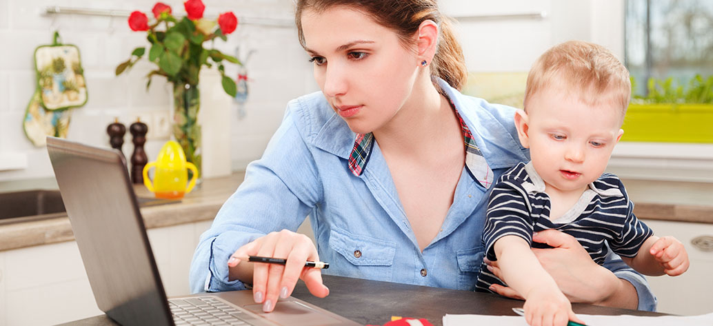 Junge Mutter mit Kind auf dem Arm füllt am Laptop den Online-Antrag zum Unterhaltsvorschuss aus 